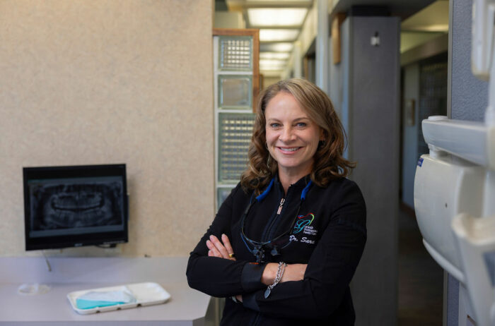 Dr. Sara Gotwalt smiling confidently in her dental office, standing with arms crossed next to dental equipment and an x-ray display in the background.